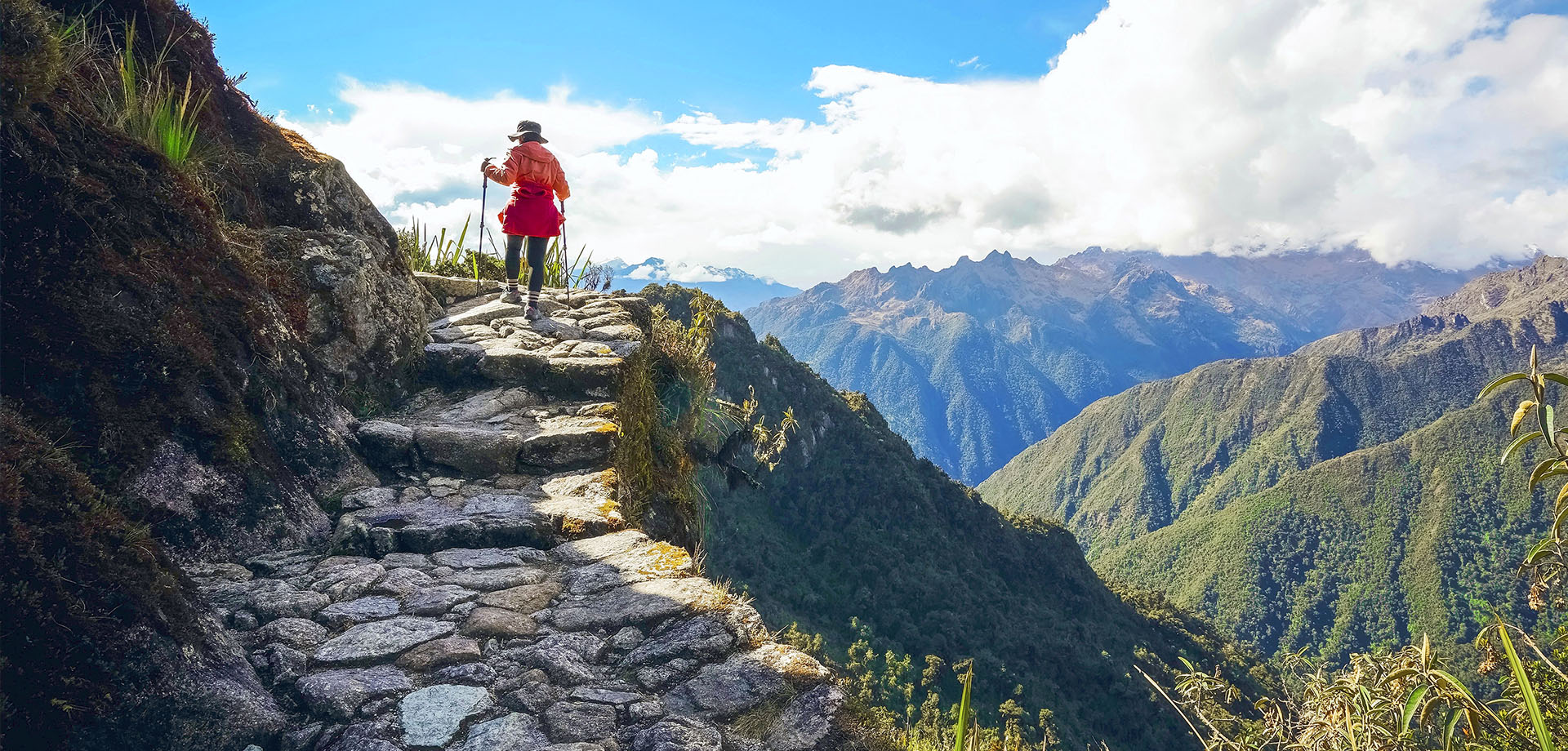 A hiker climbing the ancient stone steps of the Inca Trail with a scenic view of the Andes mountains in the background.